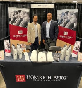 Two people stand smiling behind a booth with Homrich Berg Wealth Management banners. The table displays branded water bottles and tote bags. They are at a professional event or expo.