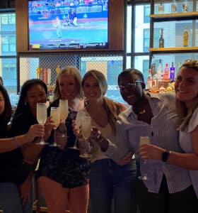 A group of six people celebrating with champagne glasses in a bar. They are smiling and standing closely together. Behind them, shelves display various bottles, and a TV shows a sports game.