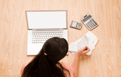A person sitting on a wooden floor is surrounded by a laptop, a smartphone, a calculator, and a pile of receipts, indicating they are working on financial or budgeting tasks.