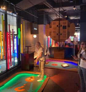 A group of people playing mini-golf indoors. One person is putting on a colorful course with glowing neon lights. Others stand nearby, watching and smiling. The background features various entertainment elements and displays.