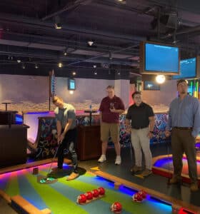 Four people standing on an indoor mini-golf course, one preparing to putt. The venue is colorful, with vibrant floor patterns and neon elements. Monitors and decorative lights are visible above.