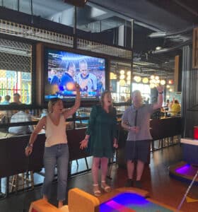 A group of people stand indoors in front of a mini-golf course, cheering with their hands raised. A TV screen above displays a sports event. The atmosphere is lively and festive.
