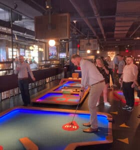 A group of people enjoying mini-golf indoors. One person is putting on a glowing, colorful course while others watch and smile. The setting is lively with neon lights and televisions in the background.