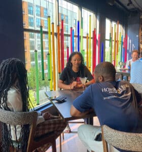A group of people sitting at a table in a modern cafe with tall, colorful glass decorations and large windows. The background shows more patrons at tables and a view of urban buildings outside.