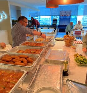 People serving themselves food from a buffet table in a bright, open room. The table is filled with trays of buns, sausages, salads, and snacks. In the background, others sit and socialize near a mini basketball game and large windows.