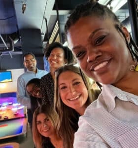 A group of six people smiling and posing together indoors. They are standing in a line, and the setting appears to be a recreational area with arcade games and tables in the background. The atmosphere is casual and friendly.