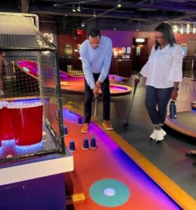 A man and woman play mini-golf indoors. The man, wearing a light blue shirt, prepares to putt while the woman, in a white top, watches. They are surrounded by colorful lights and obstacles, including large red cup props.