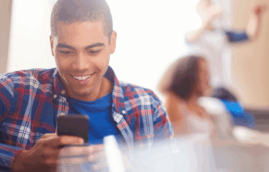 A young man in a plaid shirt smiles at his phone in a bright room. In the background, a woman is blurred and a person is raising their hands.