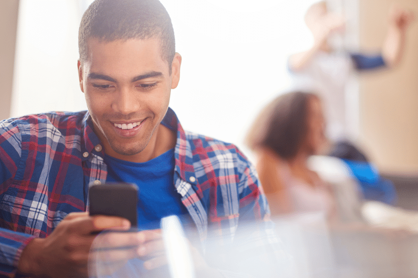 A young man in a plaid shirt smiles at his phone in a bright room. In the background, a woman is blurred and a person is raising their hands.