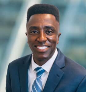 A man in a business suit and tie is smiling against a blurred indoor background.