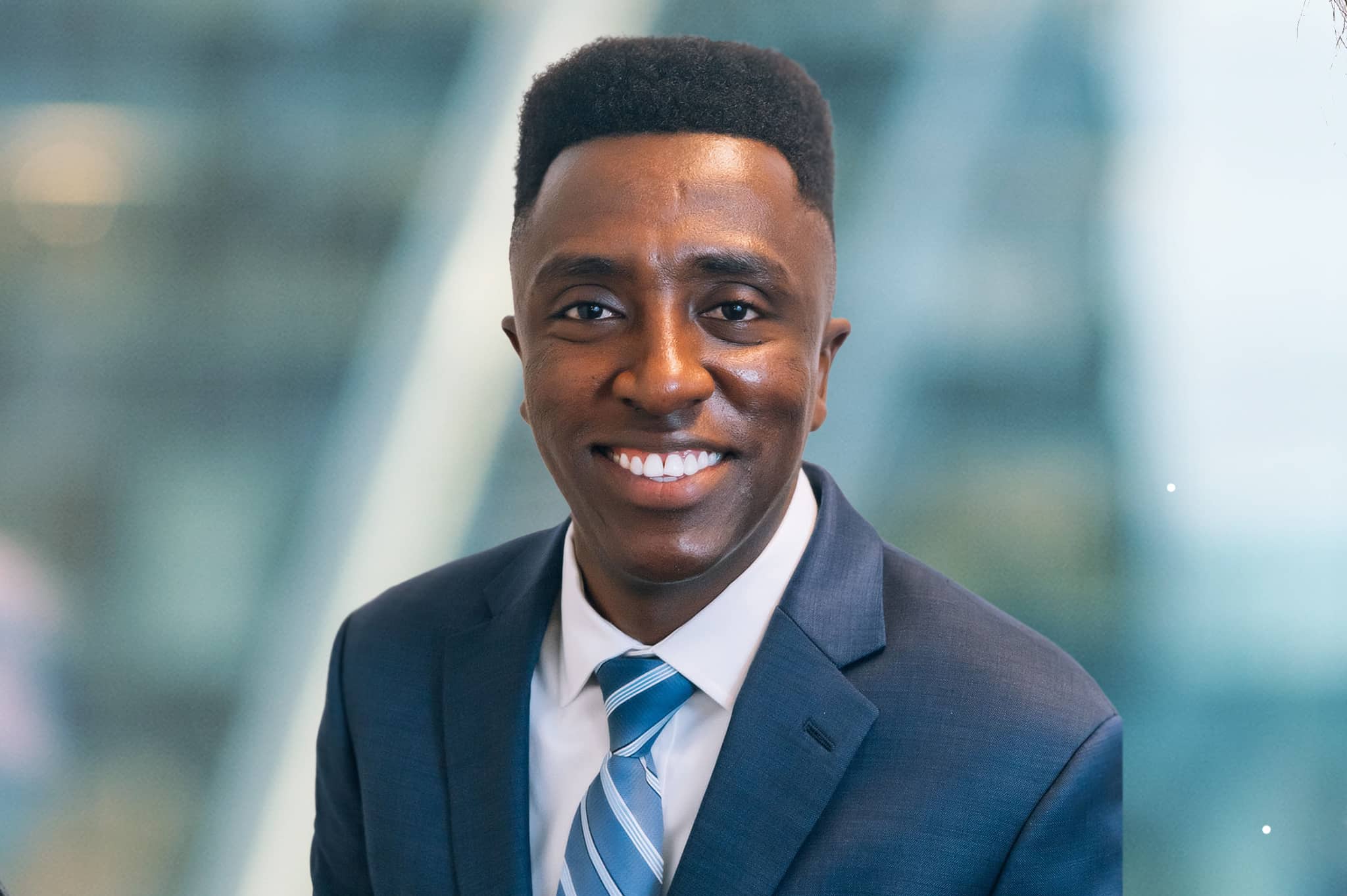 A man in a business suit and tie is smiling against a blurred indoor background.