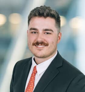 A person with short hair and a mustache is wearing a black suit and red patterned tie. They are smiling and standing against a blurred background with soft lighting.
