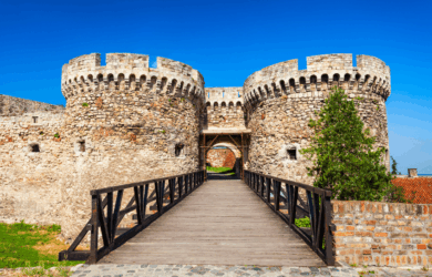 A stone fortress with two round towers and a wooden bridge under a clear blue sky. Lush greenery surrounds the structure, enhancing its historic appearance. The path leads through the towers, inviting exploration.
