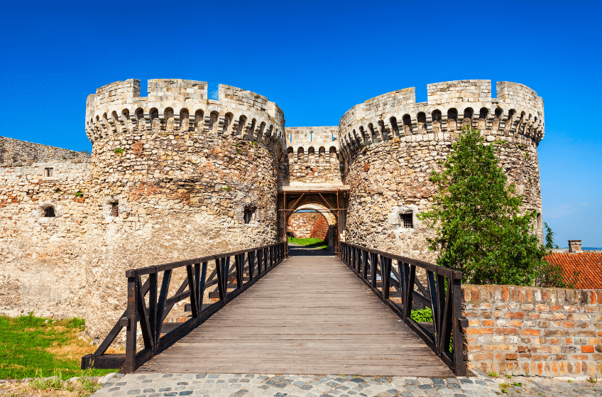 A stone fortress with two round towers and a wooden bridge under a clear blue sky. Lush greenery surrounds the structure, enhancing its historic appearance. The path leads through the towers, inviting exploration.
