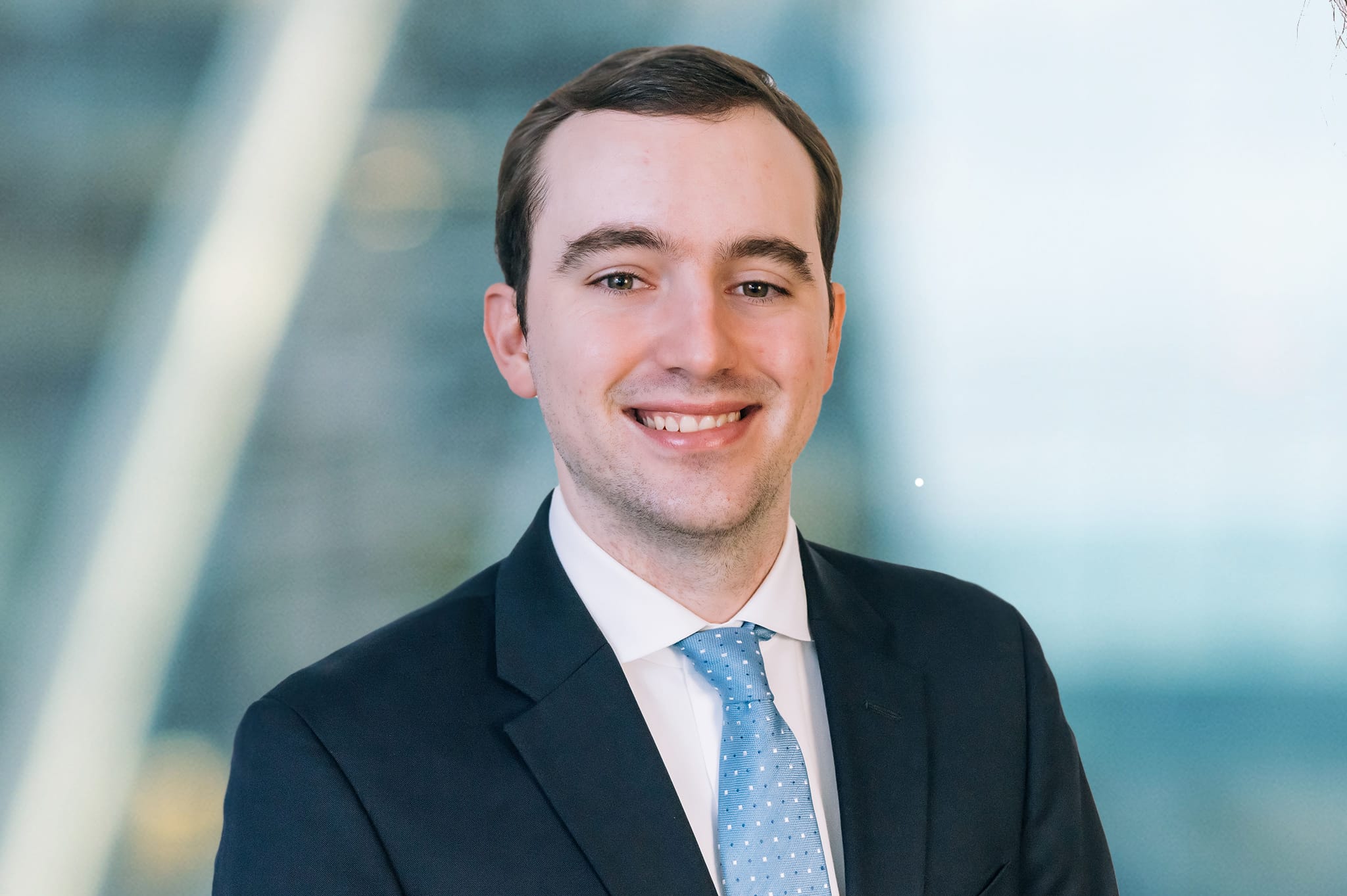 A smiling man in a business suit and blue tie stands against a blurred background. The setting appears professional with a soft, out-of-focus backdrop.