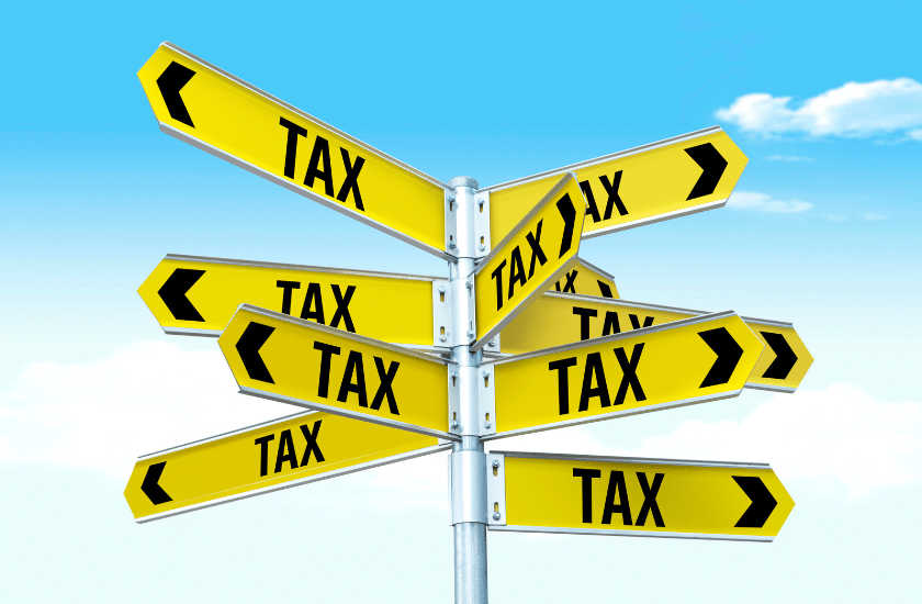A signpost with multiple yellow arrows pointing in different directions, each labeled with the word TAX in bold black letters, set against a blue sky with some clouds.