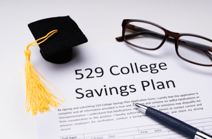 Image of a 529 College Savings Plan application form on a table. A miniature graduation cap and a pair of glasses are placed above the form, which is partially visible. A pen rests on the form, suggesting its ready to be filled out.