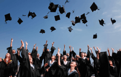 A group of graduates in black gowns and caps joyfully toss their caps into the clear blue sky, celebrating their graduation. They are looking upwards with smiles and open arms, capturing a moment of achievement and excitement.