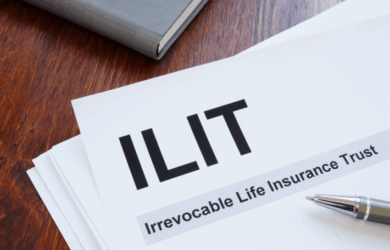 A document labeled ILIT with the subtitle Irrevocable Life Insurance Trust is on a wooden table. A pen is placed beside it, and part of a closed book or notebook is visible in the background.
