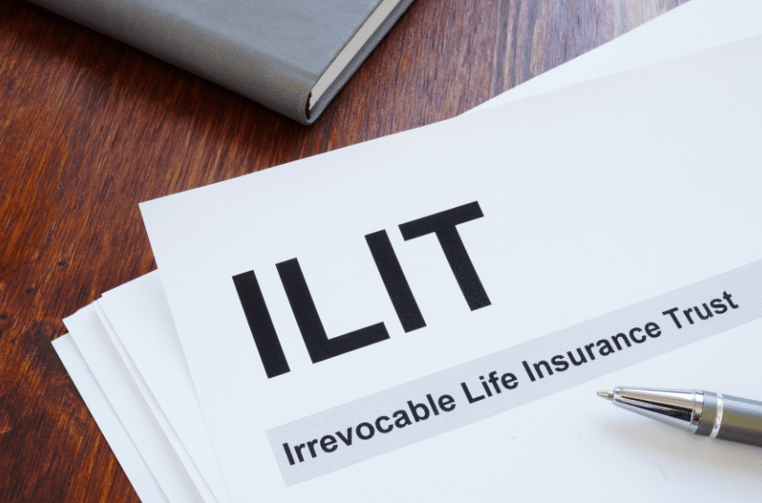 A document labeled ILIT with the subtitle Irrevocable Life Insurance Trust is on a wooden table. A pen is placed beside it, and part of a closed book or notebook is visible in the background.