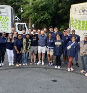 A diverse group of people posing together outdoors in front of two EcoShred trucks. Many are wearing blue shirts with I ♥️ HB on them. Trees are visible in the background. The setting appears to be informal and community-oriented.