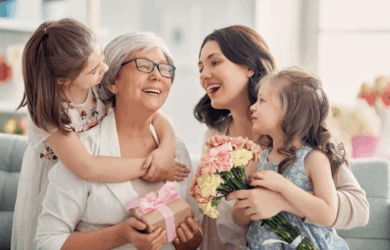 A joyful scene with a grandmother holding a wrapped gift, a woman holding a bouquet of flowers, and two young girls smiling and hugging the grandmother. Theyre gathered indoors, celebrating a happy occasion.