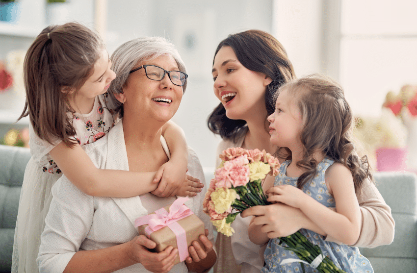 A joyful scene with a grandmother holding a wrapped gift, a woman holding a bouquet of flowers, and two young girls smiling and hugging the grandmother. Theyre gathered indoors, celebrating a happy occasion.