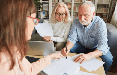 An elderly couple is sitting at a table with a woman. The man is signing a document while the woman holds papers. A laptop is open on the table, and shelves filled with books are in the background.