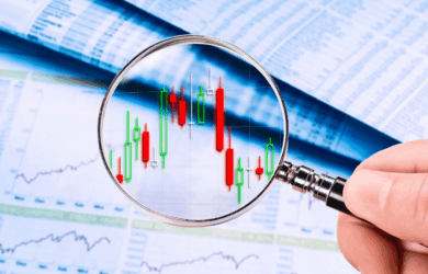 A hand holds a magnifying glass over a financial chart displaying red and green candlestick patterns. The background features blurred financial documents and graphs, suggesting analysis of stock market data.