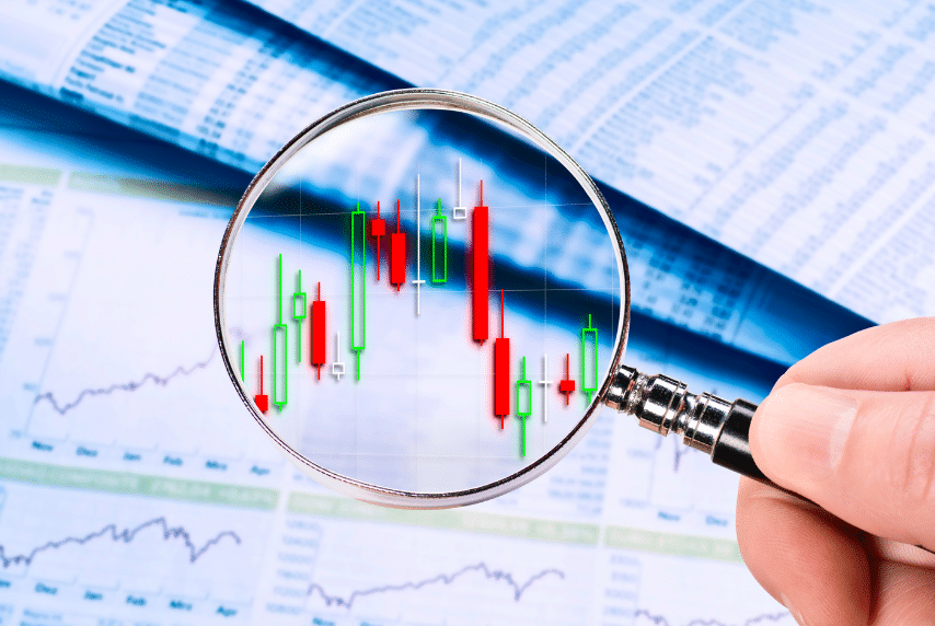 A hand holds a magnifying glass over a financial chart displaying red and green candlestick patterns. The background features blurred financial documents and graphs, suggesting analysis of stock market data.
