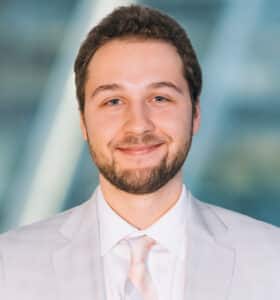 A man with short hair and a beard is smiling in a professional setting. He is wearing a light gray suit, a white shirt, and a pale pink tie. The background is softly blurred, suggesting an office or business environment.