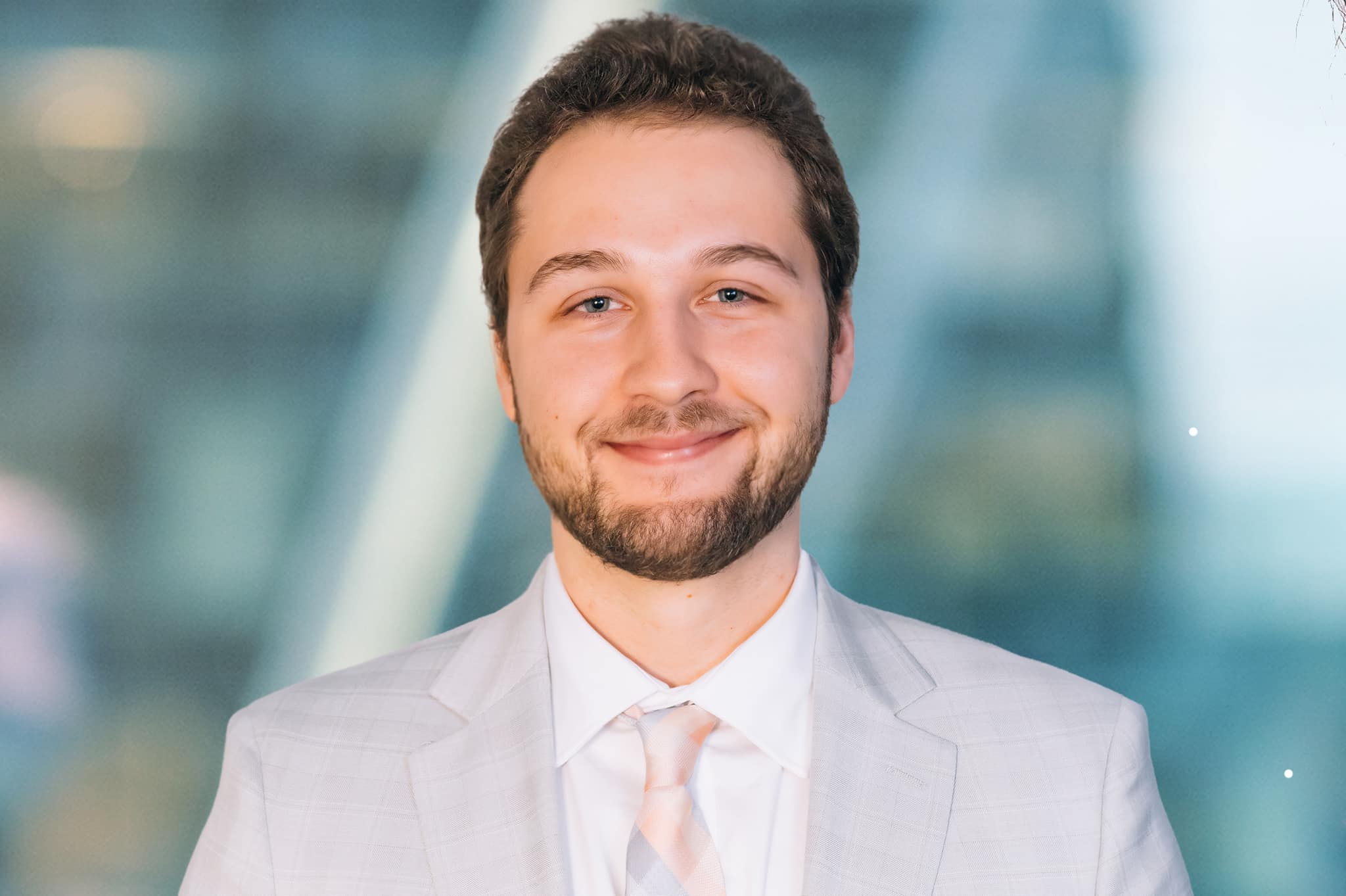 A man with short hair and a beard is smiling in a professional setting. He is wearing a light gray suit, a white shirt, and a pale pink tie. The background is softly blurred, suggesting an office or business environment.