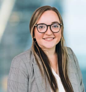 A woman with long brown hair and glasses smiles at the camera. She is wearing a gray plaid blazer and a white top. The background is softly blurred, suggesting an office or urban setting.