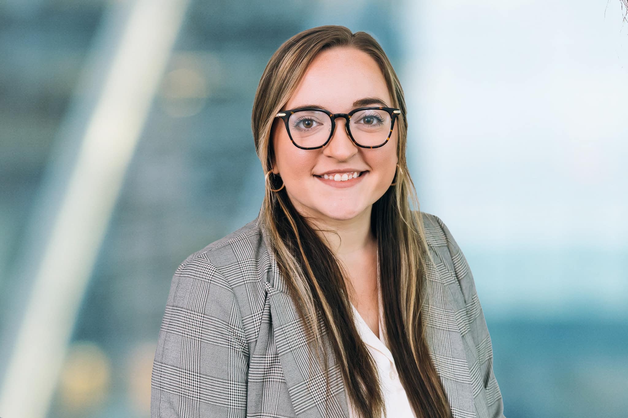 A woman with long brown hair and glasses smiles at the camera. She is wearing a gray plaid blazer and a white top. The background is softly blurred, suggesting an office or urban setting.
