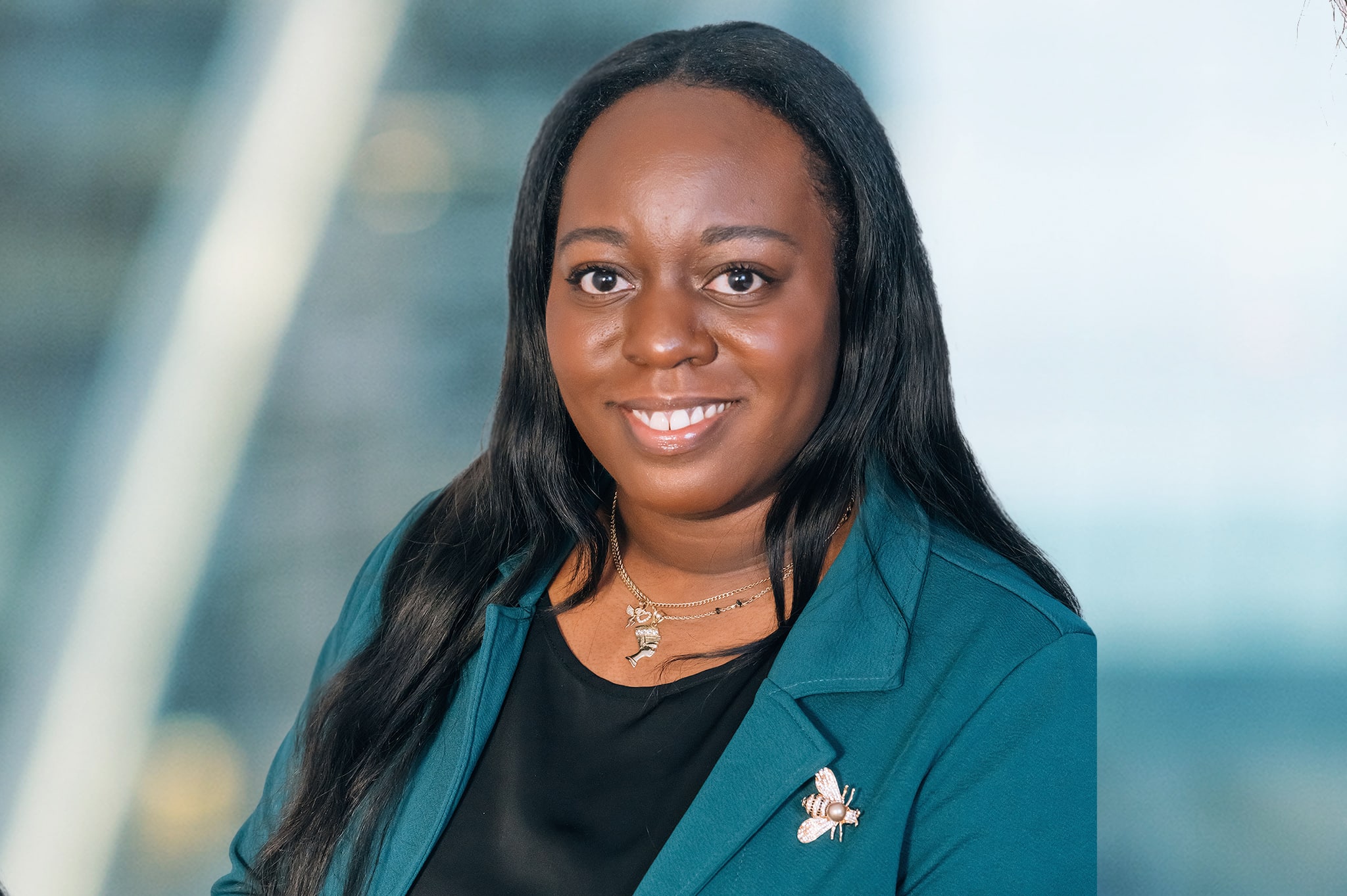 Portrait of a smiling person with long dark hair, wearing a teal blazer and black top. The background is softly blurred.