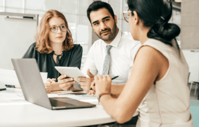 Three people sit around a table engaged in discussion. One person holds a tablet, another holds a pen, and a laptop is open on the table. They appear focused on their conversation in an office setting.