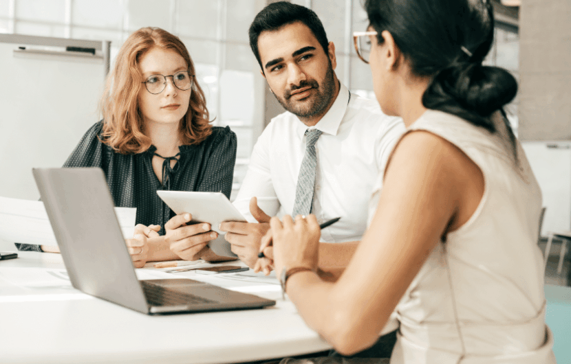 Three people sit around a table engaged in discussion. One person holds a tablet, another holds a pen, and a laptop is open on the table. They appear focused on their conversation in an office setting.