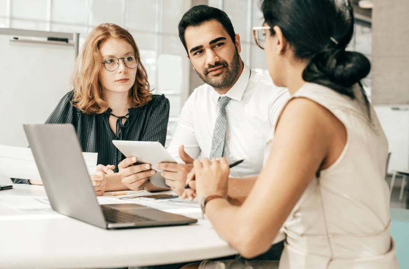 Three people sit around a table engaged in discussion. One person holds a tablet, another holds a pen, and a laptop is open on the table. They appear focused on their conversation in an office setting.