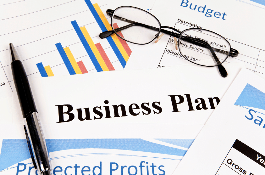 An image showing a business plan document surrounded by graphs and financial documents, with a pair of glasses and a pen resting on top. The charts display colorful bar graphs and scattered budget figures.