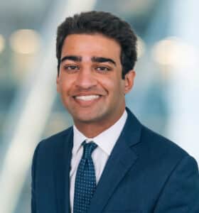 A man in a dark suit and tie smiles warmly at the camera. The background is softly blurred with a mix of neutral colors, suggesting a professional indoor setting.