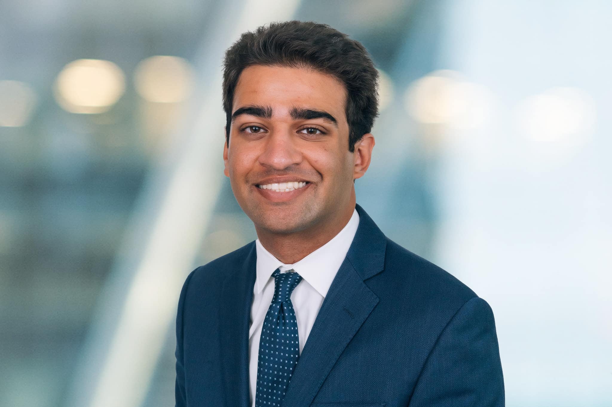 A man in a dark suit and tie smiles warmly at the camera. The background is softly blurred with a mix of neutral colors, suggesting a professional indoor setting.