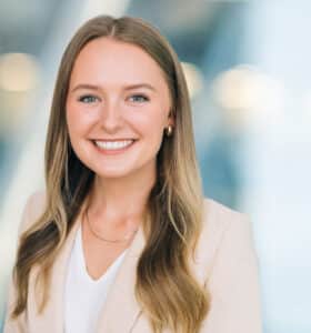 A woman with long blonde hair is smiling at the camera. She is wearing a light-colored blazer over a white top. The background is softly blurred, featuring some light streaks and round light spots.