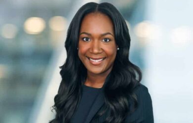 A person with long, wavy hair smiles warmly at the camera. They are wearing a dark blazer. The background is softly blurred, highlighting their friendly expression.