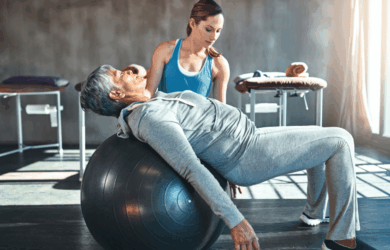 A senior woman exercises on a stability ball while a trainer assists her. The woman is wearing a gray tracksuit, and the trainer is in a blue tank top. Sunlight streams through a window, illuminating the gym setting.