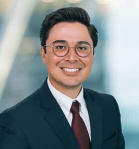 A person with short dark hair and glasses is smiling, wearing a suit, white shirt, and dark red tie. The background is blurred, suggesting an office environment.