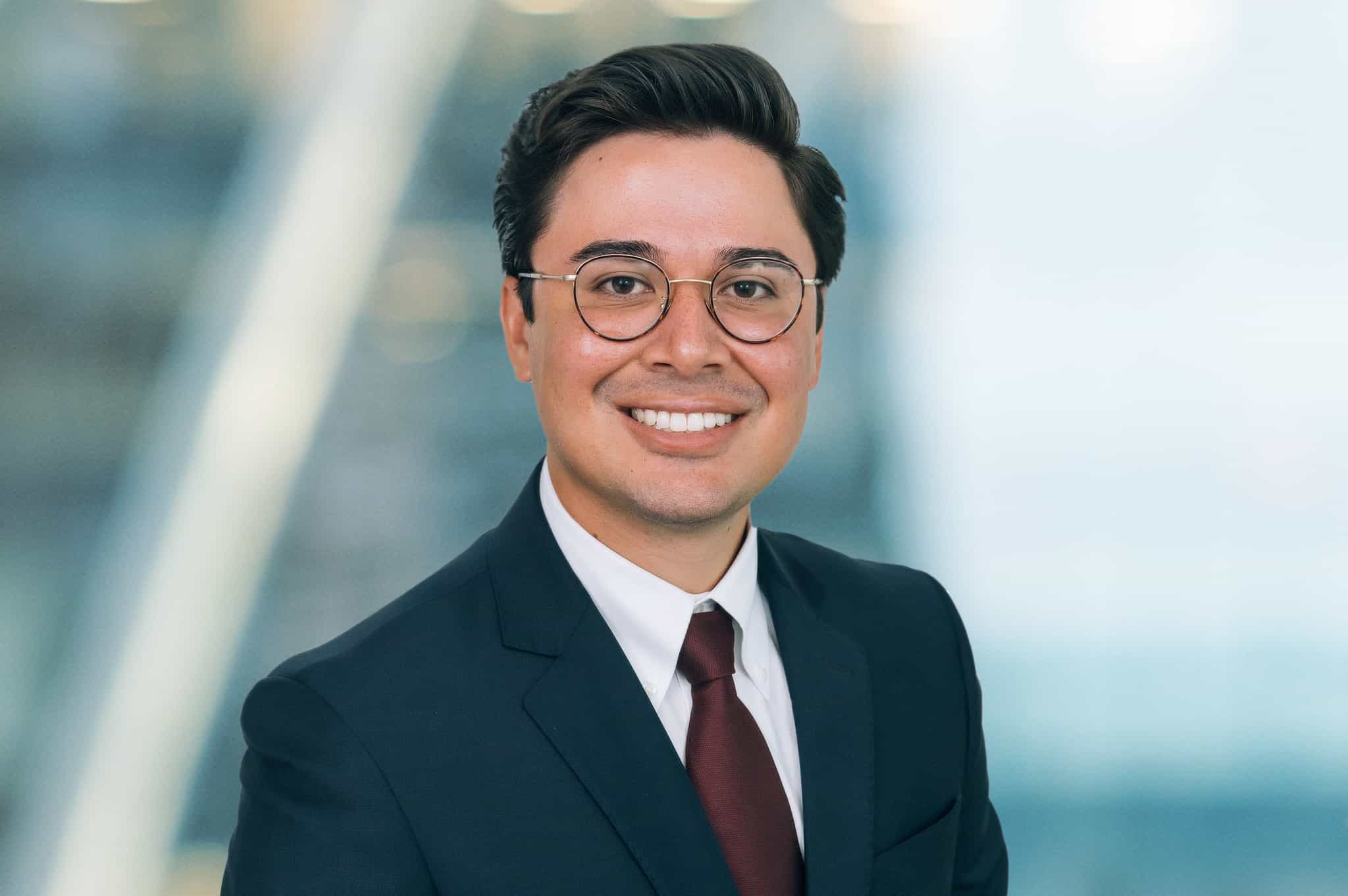 A person with short dark hair and glasses is smiling, wearing a suit, white shirt, and dark red tie. The background is blurred, suggesting an office environment.