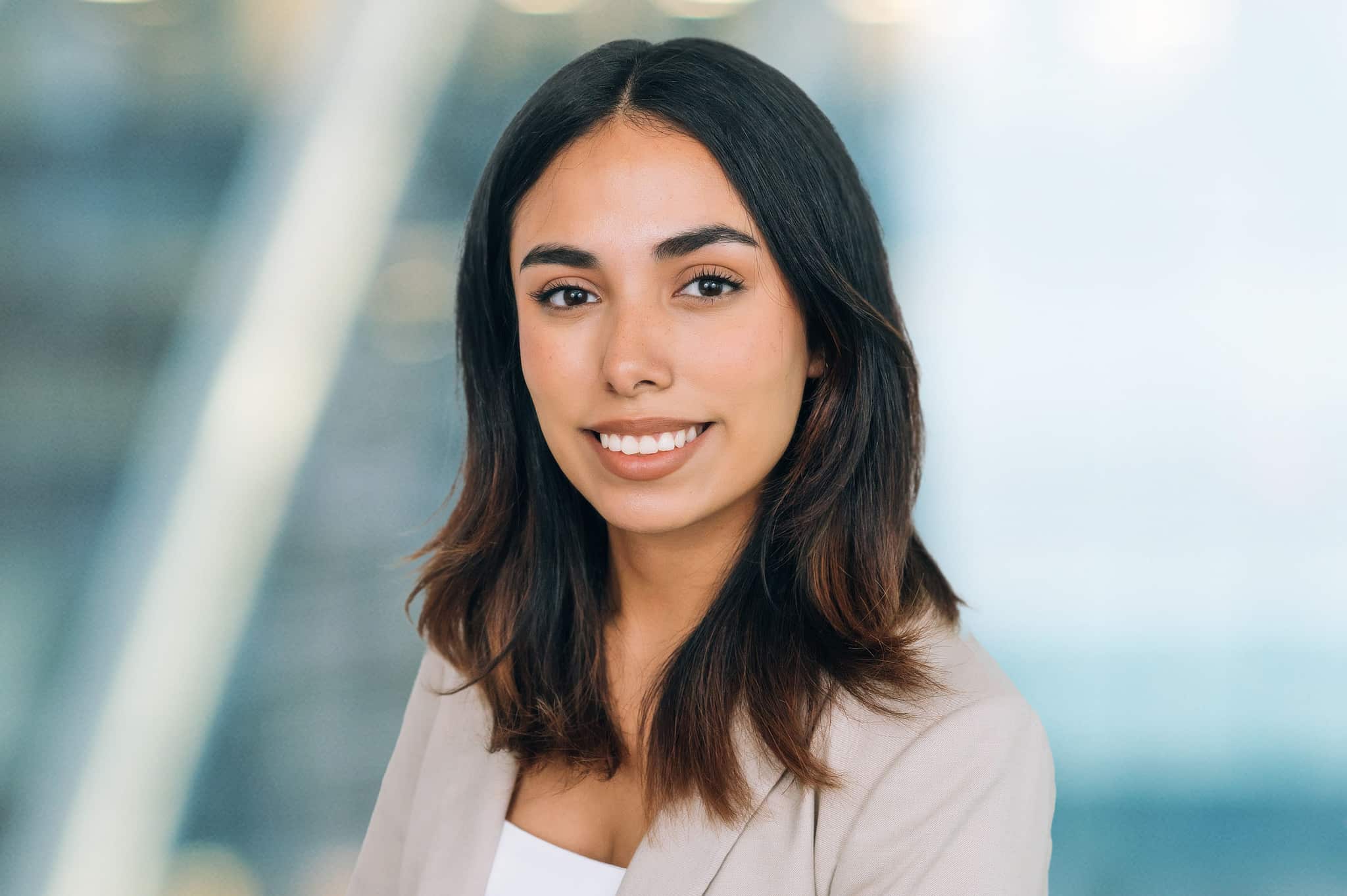 A woman with long dark hair and a beige blazer smiles, standing against a blurred background.