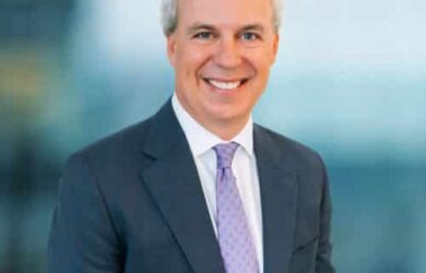 A man in a dark suit and light purple tie smiles at the camera. He has short, light-colored hair. The background is blurred, creating a professional, yet approachable atmosphere.