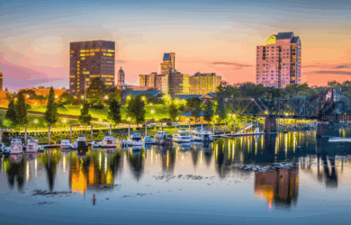 A picturesque cityscape at sunset with tall buildings, a bridge, and a waterfront. The sky is a gradient of orange and blue, and trees line the riverbank. Boats are docked in the water, reflecting the city lights.