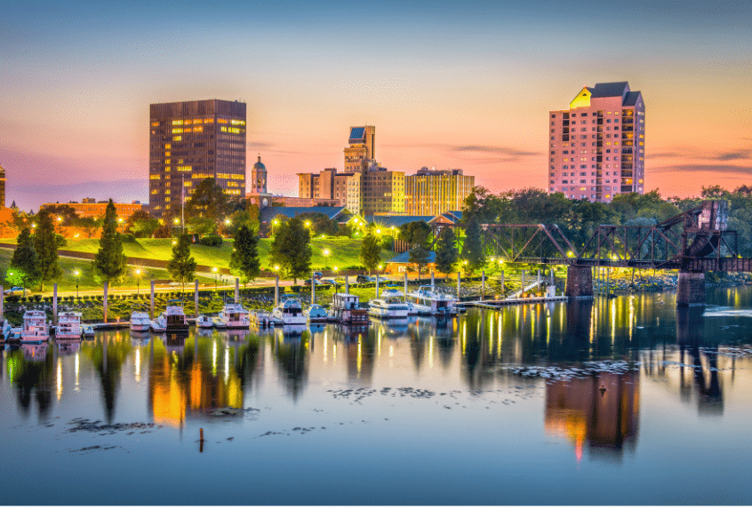 A picturesque cityscape at sunset with tall buildings, a bridge, and a waterfront. The sky is a gradient of orange and blue, and trees line the riverbank. Boats are docked in the water, reflecting the city lights.